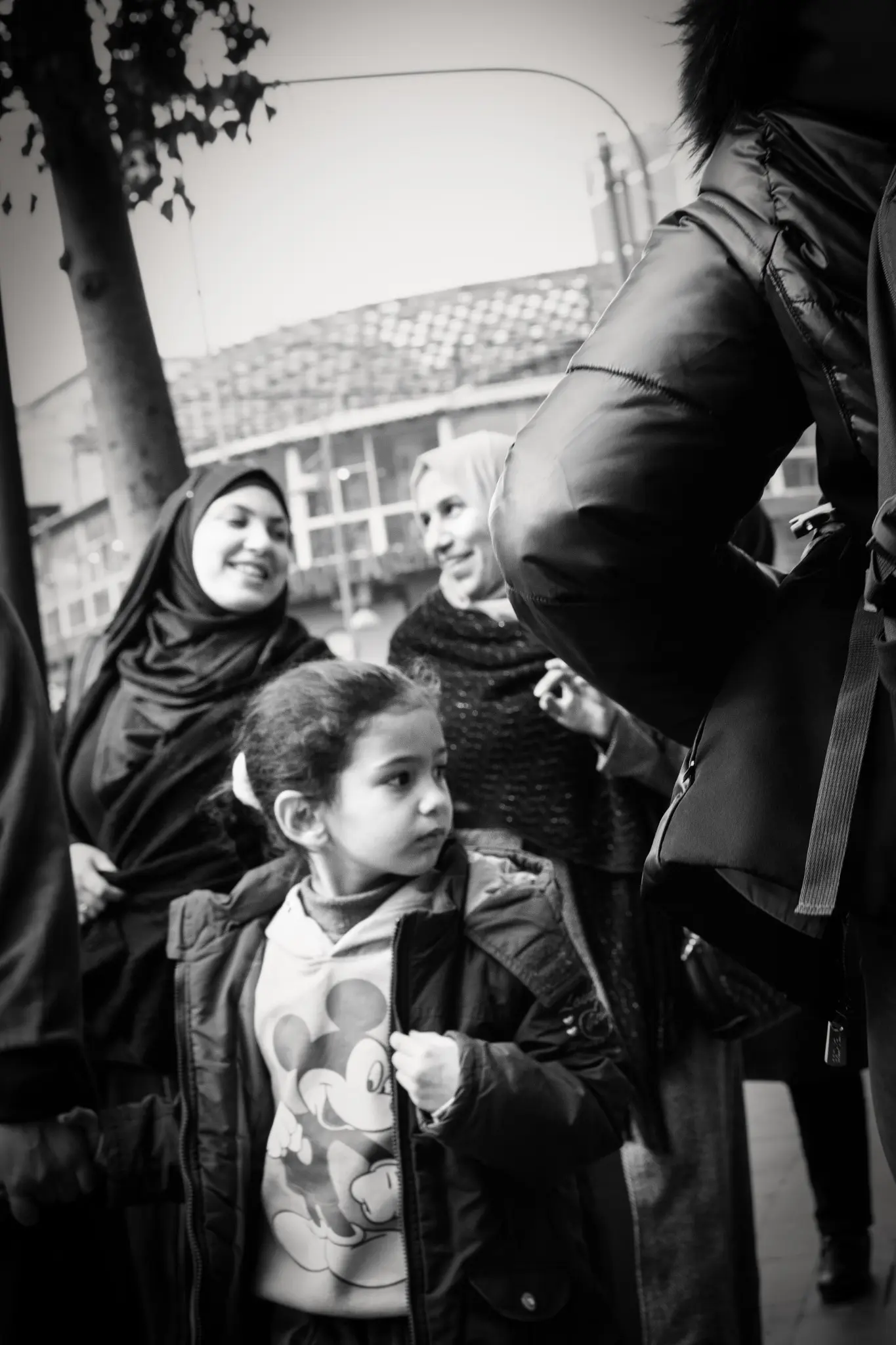 A girl watches passers-by while two women smile behind her.
