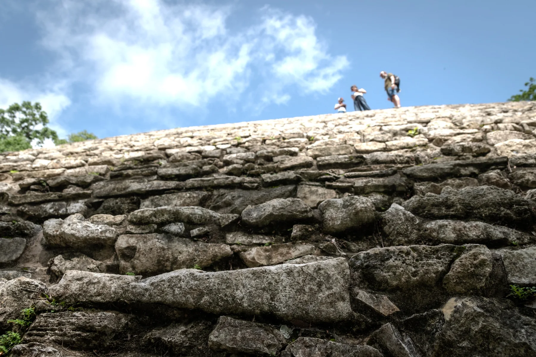 Maya ruins seen from below, with three people standing along the upper edge.