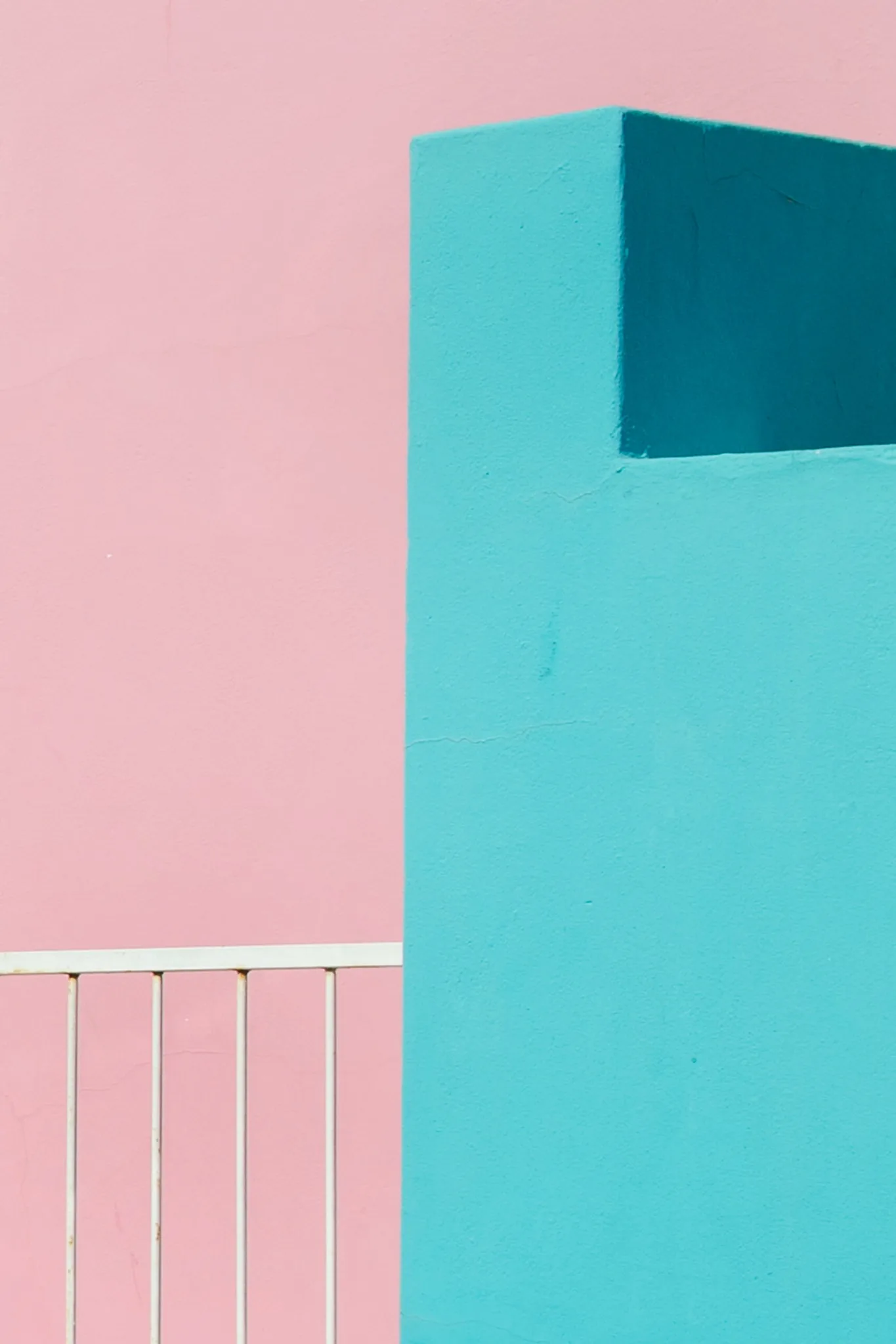 A geometric turquoise volume against a pink background with a white railing. A geometric turquoise volume against a pink background with a white railing.
